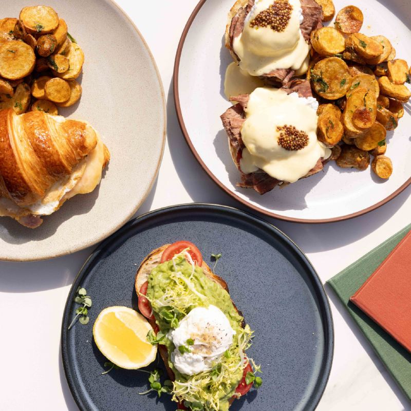 Plates of brunch dishes including avocado toast with poached egg, a croissant sandwich with potatoes, and eggs Benedict with roasted potatoes.