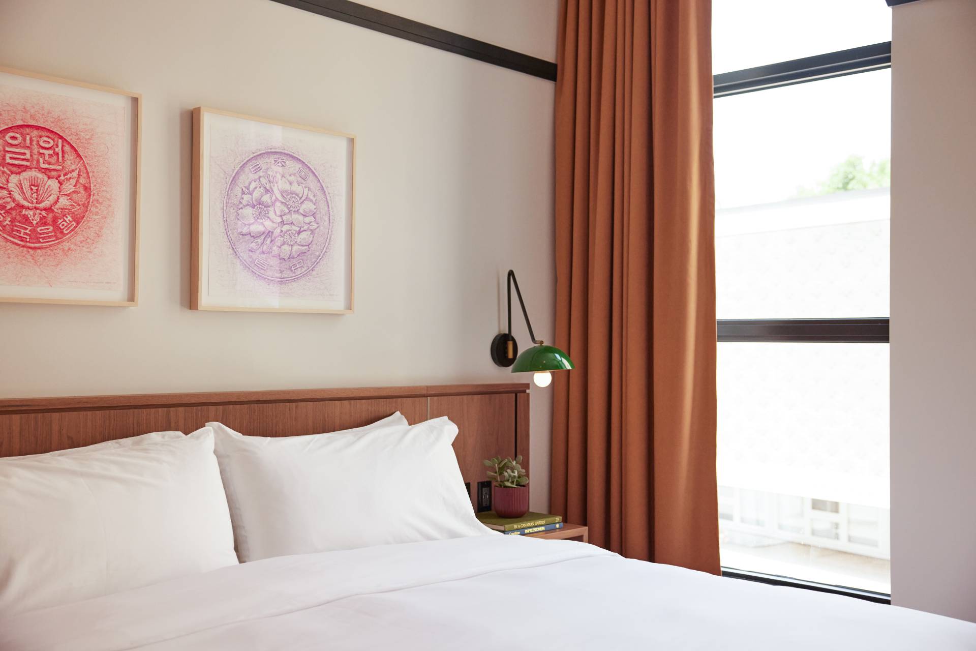 Modern hotel bedroom with white bedding, wood accents, framed wall art, rust-coloured curtains, and a large window.